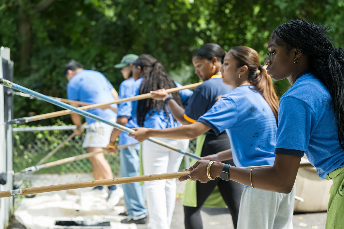 University of Rochester students in blue shirts collaboratively painting a fence during Wilson Community Service Day.
